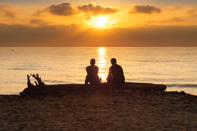 Rear view of male friends sitting at beach against orange sky