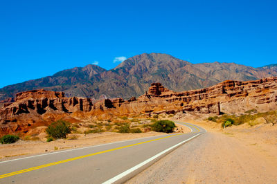 Road leading towards mountains against clear blue sky