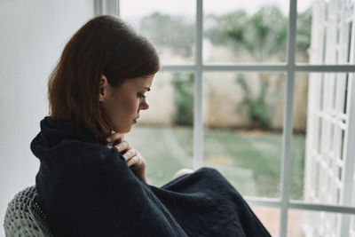 Side view of young woman looking through window at home