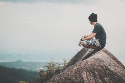 Man sitting on rock looking at mountain against sky