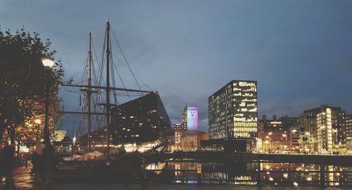 Illuminated modern buildings by river against sky at dusk