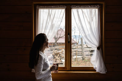 Woman sitting at the window of a mountain cabin, enjoying a cup of tea and the view.