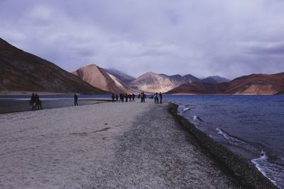 People at beach by mountains against cloudy sky