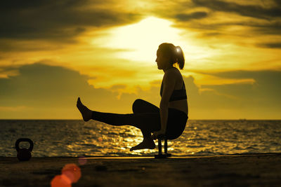 Woman exercising with dumbbells at beach against sky during sunset