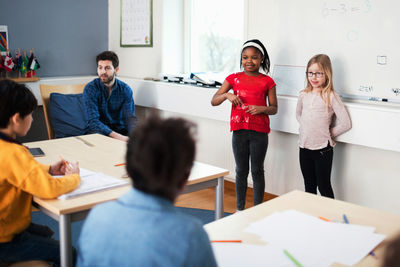 Female students standing against whiteboard wile teacher sitting in classroom