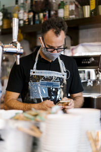 Male barista in protective face mask using smart phone at cafe during covid-19