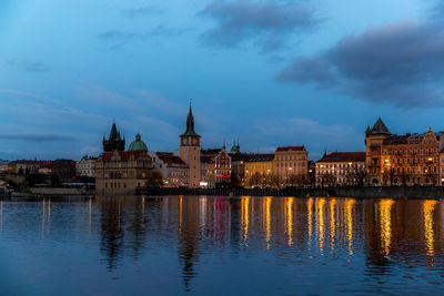 Illuminated buildings at waterfront against cloudy sky