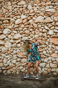 Young beautiful girl riding a skateboard in front of a stone wall
