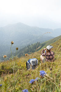 Woman traveler sitting on meadow in mountain next to backpack drinking water from bottle hiking