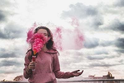 Low angle view of young woman holding smoke bomb while standing against cloudy sky