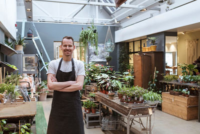 Portrait of smiling owner standing with arms crossed against potted plants at store