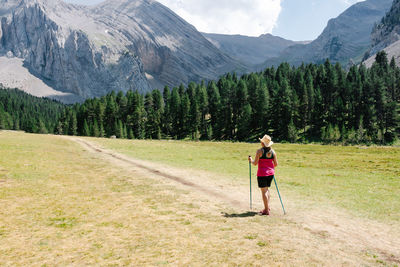 Hiker woman wearing straw hat, shorts and backpack on the path across a plain walking with amazing mountains at the background while enjoys the natural environment around. horizontal photo.