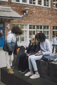 Friends sharing smart phone while sitting on retaining wall in school campus