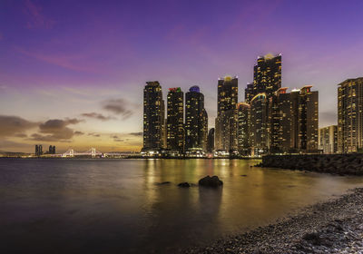 Illuminated city by river against sky at sunset