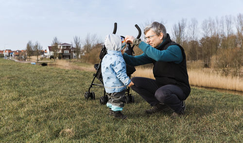 Grandfather with granddaughter crouching on grassy field against sky