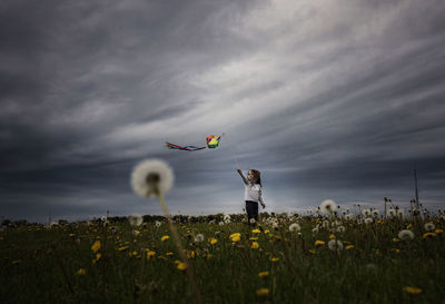 Girl playing with kite while standing on dandelion field against stormy clouds