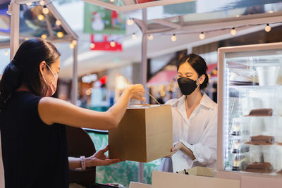 Asian woman owner bakery store wears face mask giving takeaway food bag to customer