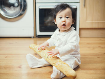 Cute baby girl sitting with baguette on hardwood floor at home