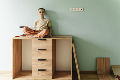 Smiling boy sitting on cabinet in front of wall at home