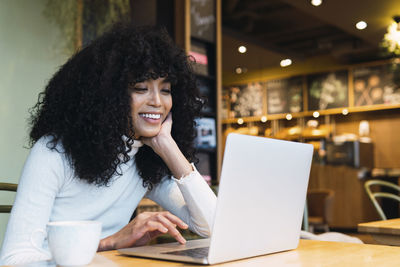 Smiling young woman with hand on chin using laptop at cafe