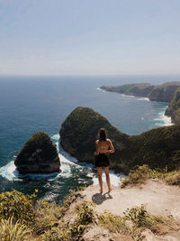 Rear view of woman looking at sea while standing on mountain against sky during sunny day