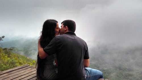 Rear view of young couple kissing while sitting on platform against fog