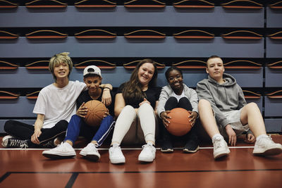 Portrait of smiling male and female friends sitting with basketballs in sports court