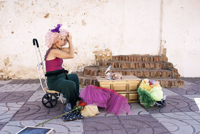 Side view of concentrate middle aged female clown with bright makeup in costume putting on wig and looking away while sitting on circus chair with wheels on street