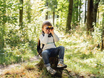 Little explorer on hike in forest. boy with binoculars sits on stump.outdoor activity for children. 