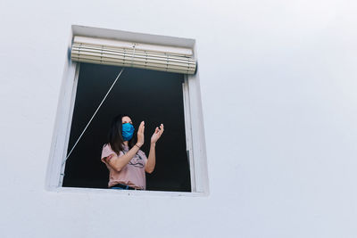 Young brunette woman with mask clapping from the window of the house with white background