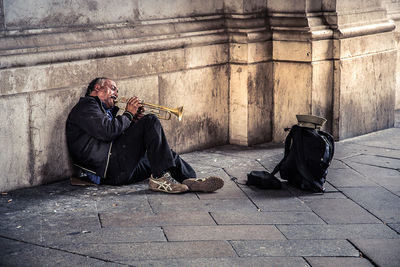 Senior street musician playing musical instrument while sitting on street