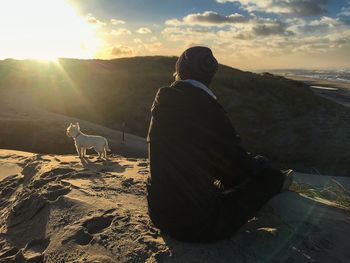 Rear view of man with dog sitting on landscape against sky during sunset