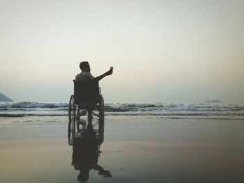 Man taking selfie while sitting on wheelchair at beach during sunset