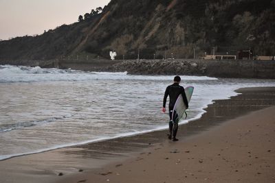 Rear view of man with surfboard walking at beach against mountain during sunset