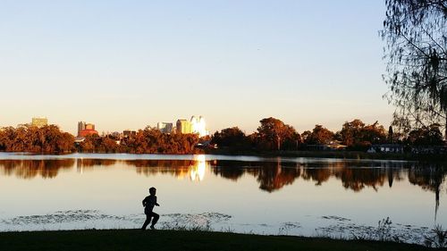 Silhouette boy standing by lake against clear sky during sunset