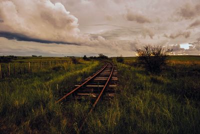 Railroad track on grassy field against cloudy sky during sunset