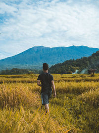 Rear view of man walking on grassy field against cloudy sky