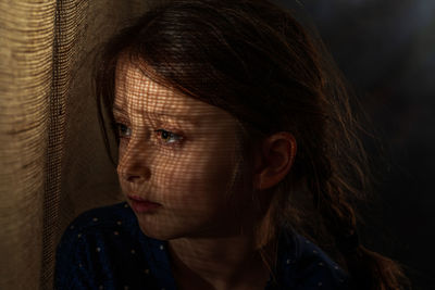 Close-up girl with shadow on face looking away while standing by burlap at darkroom