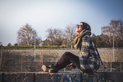 Smiling young woman sitting on retaining wall against clear sky