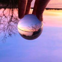 Cropped hand holding crystal ball with reflection of lake against sky during sunset