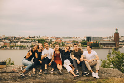 Full length of happy friends sitting on rock formation against city