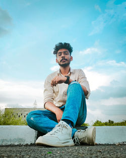 Young man sitting on retaining against sky