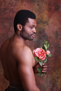 Shirtless young man holding flower while standing against wall