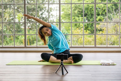 Smiling woman filming while doing stretching exercise through smart phone on tripod at yoga studio