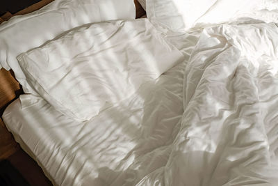 Unmade white bed with white bed sheet and pillow in the bedroom in the morning with sun and shadow