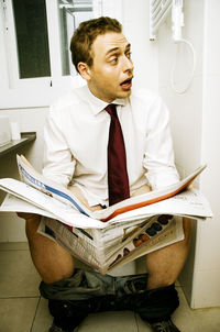 Shocked young man reading newspaper on toilet seat