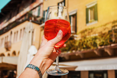 Female hand with glass of orange cocktail spritz near old buildings. sunny day in verona, italy