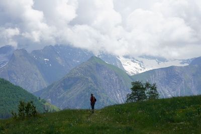 Hiker standing on grassy field by mountains against cloudy sky