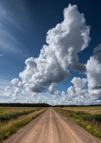 Dirt road amidst green field against cloudy sky during sunny day