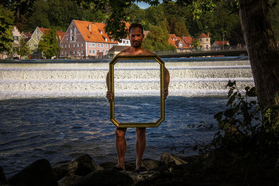 Portrait of shirtless man holding mirror while standing against river